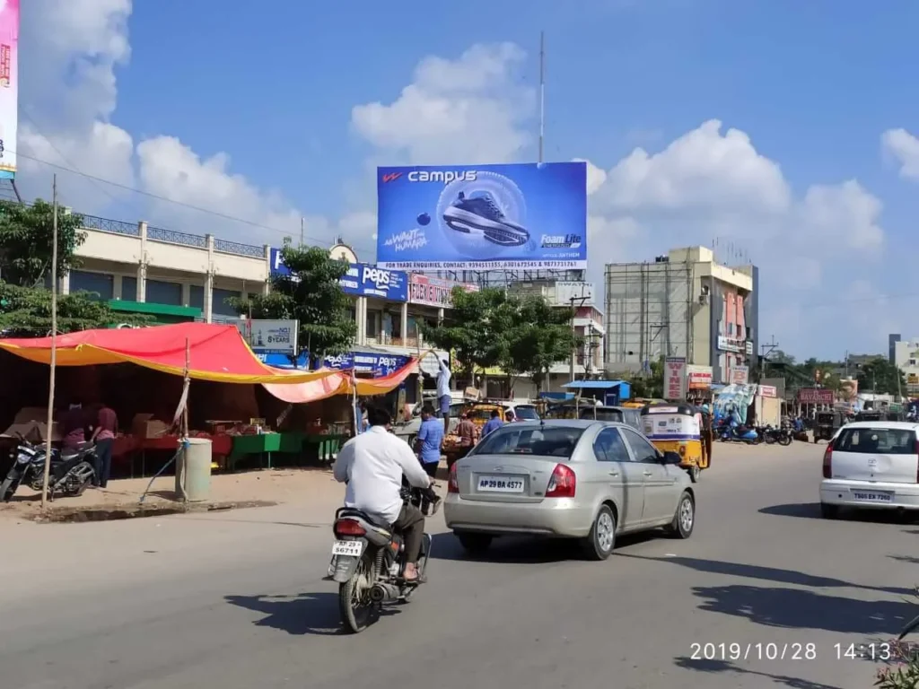 A busy street in India featuring a large, colorful hoarding advertisement for Plastivision, a plastics exhibition. The hoarding is prominently placed above the road, ensuring high visibility for passing vehicles, including cars, motorcycles, and auto-rickshaws. Trees and buildings surround the scene, adding depth to the urban setting.