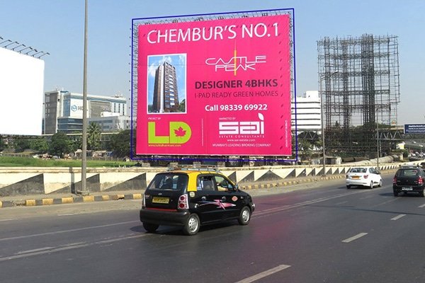 A busy street in India featuring a large, colorful hoarding advertisement for Plastivision, a plastics exhibition. The hoarding is prominently placed above the road, ensuring high visibility for passing vehicles, including cars, motorcycles, and auto-rickshaws. Trees and buildings surround the scene, adding depth to the urban setting.