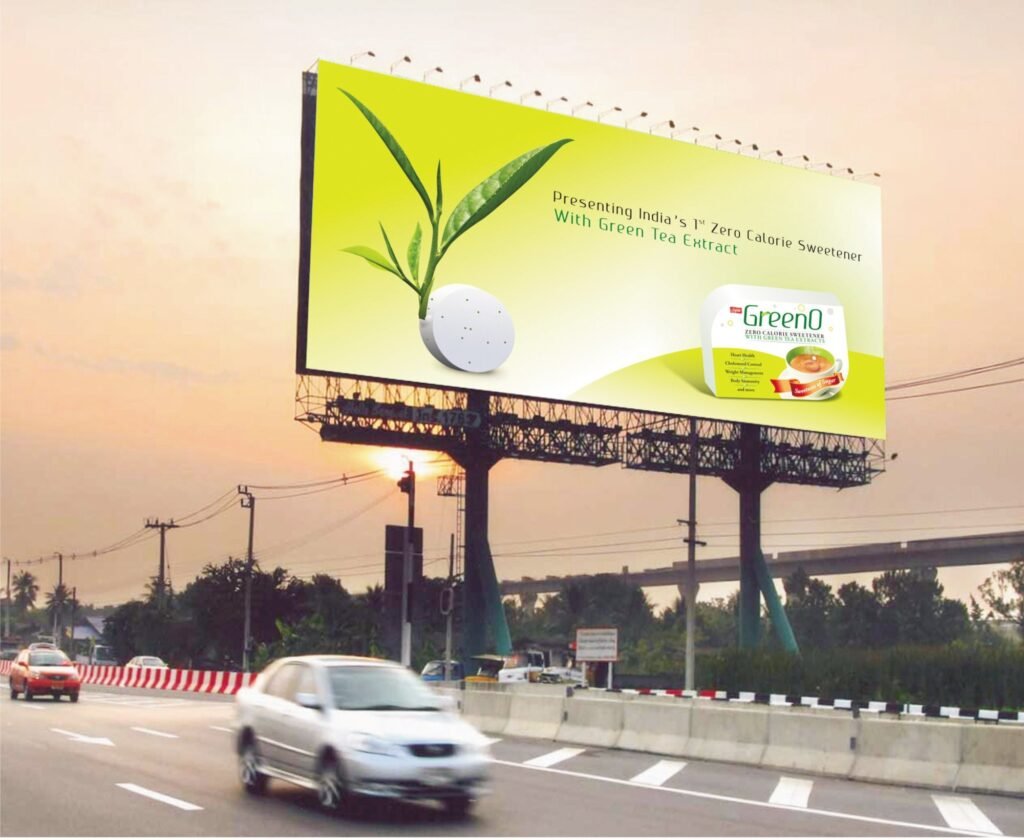 A busy street in India featuring a large, colorful hoarding advertisement for Plastivision, a plastics exhibition. The hoarding is prominently placed above the road, ensuring high visibility for passing vehicles, including cars, motorcycles, and auto-rickshaws. Trees and buildings surround the scene, adding depth to the urban setting.