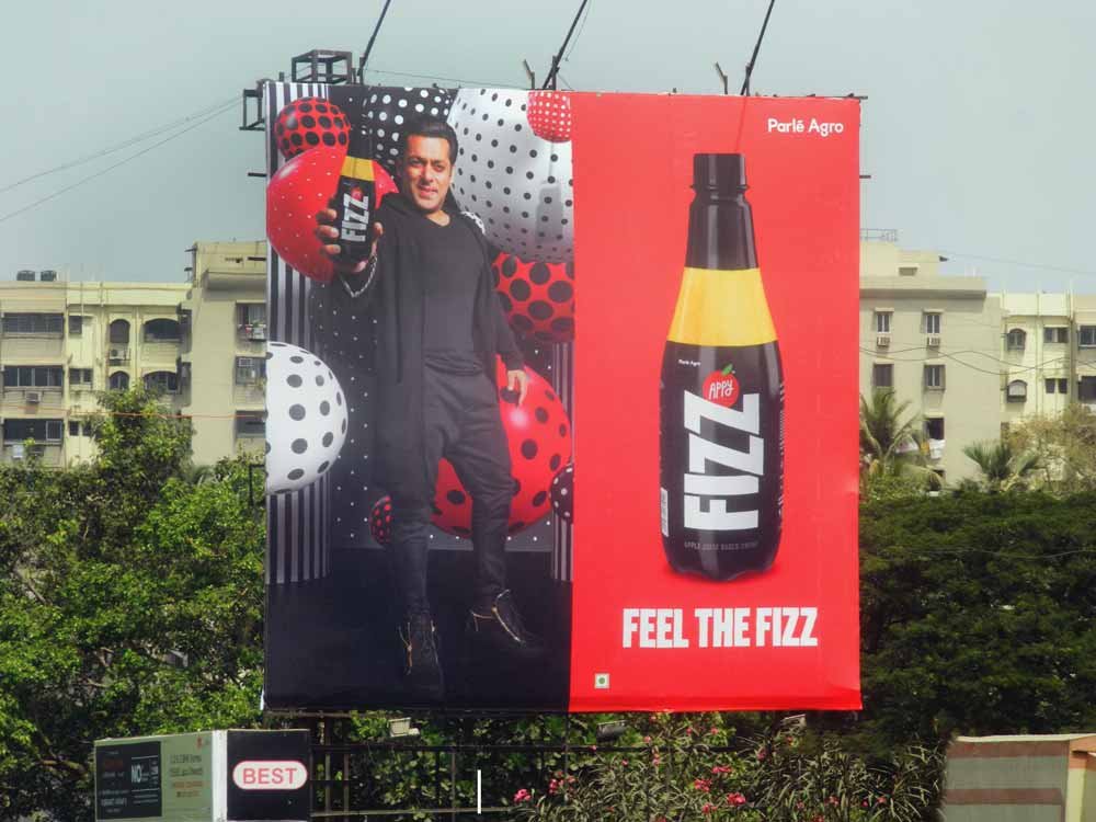 A busy street in India featuring a large, colorful hoarding advertisement for Plastivision, a plastics exhibition. The hoarding is prominently placed above the road, ensuring high visibility for passing vehicles, including cars, motorcycles, and auto-rickshaws. Trees and buildings surround the scene, adding depth to the urban setting.